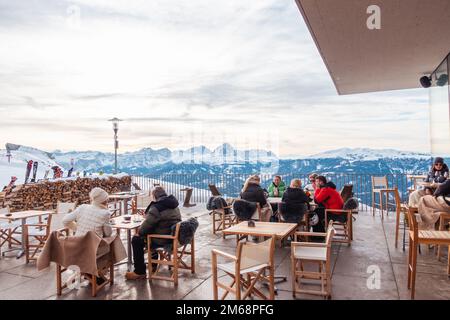 Eine Gruppe von Touristen sitzt an den Tischen eines Cafés hoch oben in den Alpen am Kronplatz (Plan de Coroes) mit schneebedeckten Alpen im Hintergrund. Touristen Stockfoto