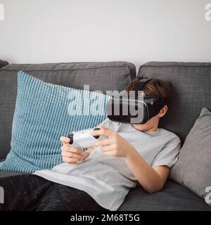 Männliches Kind aus Kaukasien, das ein VR-Headset trägt und ein Gamepad hält, während es im Wohnzimmer sitzt und Virtual-Reality-Simulationsspiele spielt Stockfoto