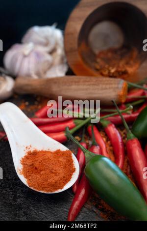 Chilipulver auf einem weißen Löffel mit einem Holzpistill im Hintergrund, umgeben von Knoblauch mit rotem und grünem Chilis zusammen mit Chiliflocken Stockfoto