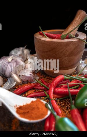 Chilipulver auf einem weißen Löffel mit einem Holzpistill im Hintergrund, umgeben von Knoblauch mit rotem und grünem Chillis Stockfoto