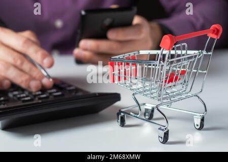 Online-Shopping-Konzept. Kleiner Supermarkt, Einkaufswagen zum Einkaufen, Ein Mann zählt die Kosten für Einkäufe mit einem Taschenrechner. Stockfoto