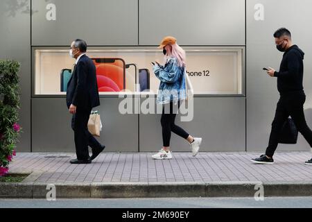 TOKIO, JAPAN - 8. April 2021: Fußgänger, die Smartphones verwenden, passieren einen Apple Store in Tokios Ginza-Gegend mit einem Werbespot für das iPhone 12. Stockfoto