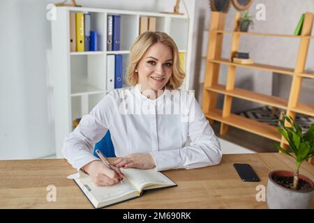 Porträt einer glücklichen Geschäftsfrau, die sich Notizen macht, während sie im Büro am Tisch sitzt. Stockfoto