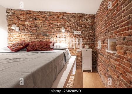 Schlafzimmer eines freiliegenden Loft-Apartments aus Ziegelstein mit einem Doppelbett mit roten Kissen und einem weißen Nachttisch aus Holz Stockfoto