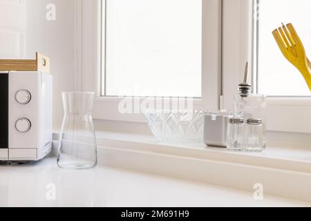 Ecke einer Küche mit einer synthetischen Arbeitsplatte mit weißen Aluminiumfenstern, Glasgefäßen und einer Mikrowelle Stockfoto