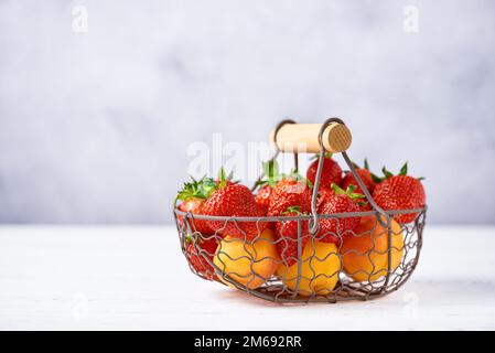 Reife Erdbeeren und Aprikosen in einem Korb aus Metall auf einem weißen Holztisch. Kopierraum, Nahaufnahme. Stockfoto