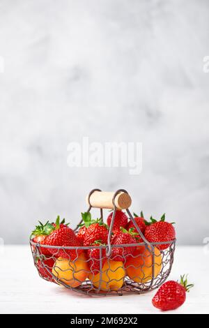 Frische Erdbeeren und Aprikosen in einem Korb aus Metall auf einem weißen Holztisch. Grauer Hintergrund. Vertikal, Kopierraum, Nahaufnahme. Stockfoto