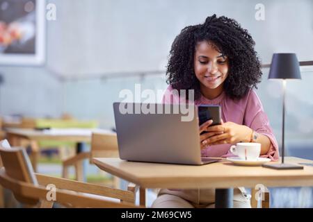 Porträt einer Geschäftsfrau, die lässig angezogen ist und auf das Telefon schaut, während sie in einem Café sitzt und arbeitet. Stockfoto