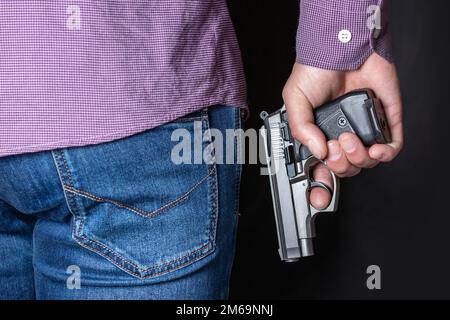 Ein Mann, der hinter seinem Rücken eine Waffe in der Hand hält, Nahaufnahme. Konzepte: Verbrechen, versuchter Mord, eine Schusswunde, der Mörder, Raub Stockfoto