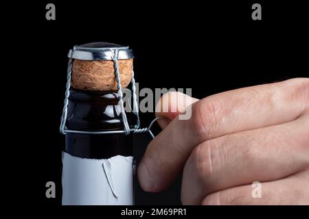 Die Hand des Mannes öffnet eine Flasche Champagner. Der Mann dreht das Champagnermuselett ab. Champagnerflasche auf schwarzem Hintergrund Stockfoto