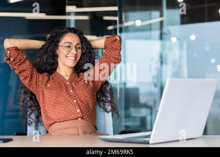 Glückliche und lächelnde erfolgreiche hispanische Geschäftsfrau beendete Arbeit zufrieden mit Arbeitsergebnis und Leistung, Hände hinter dem Kopf und lächelnd aus nächster Nähe. Stockfoto