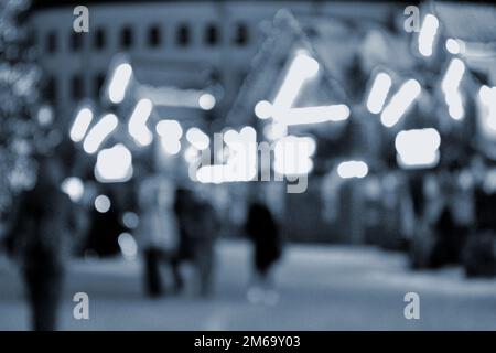 Unscharfer Hintergrund. In der Winternacht laufen die Leute in der Stadt herum. Schwarze Silhouetten von Menschen, die in der Nähe von Häusern spazieren, dekorierte, leuchtende Beleuchtung. Weiße Bokeh-Spots von leuchtenden Hauslichtern Blaue Farbe Stockfoto