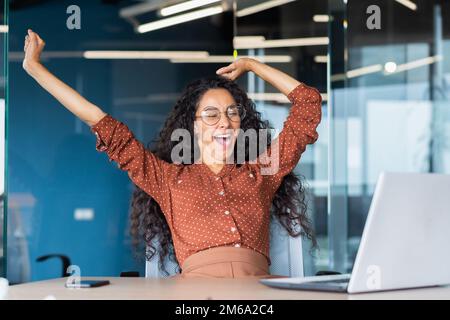 Müde Geschäftsfrau im Büro, die sich streckt und gähnt, lateinamerikanische Frau, die im Innenbereich am Arbeitsplatz arbeitet, mit einem gelangweilten Laptop übermüdet. Stockfoto