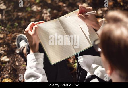 Aus einem großen Blickwinkel geöffnetes Notizbuch mit handschriftlichem Text, Person, die im Park auf dem Boden sitzt und Notizen im Tagebuch schreibt. Stockfoto