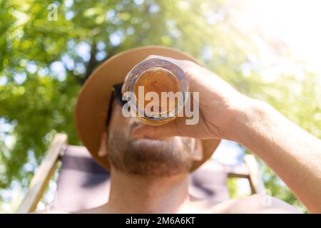 Erwachsener Mann mit Strohhut trinkt Bier im Sommer mit Blick aus dem unteren Winkel. Stockfoto