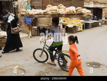 Ein kleiner Junge, der ein Tablett Pitta-Brot auf seinem Kopf mit dem Fahrrad bringt - Kairo, Ägypten Stockfoto