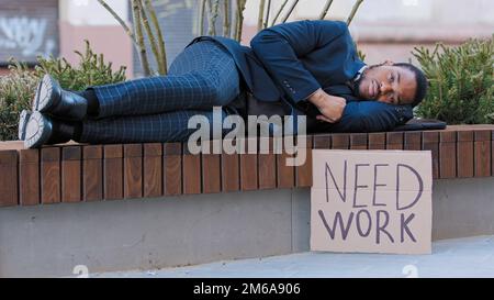 Schlafende Obdachlose gefeuerte afroamerikanische Männer schlafen auf einer Bank in der Stadt mit Poster brauchen Arbeit. Ein einsamer, aufgebrachter, ethnischer Typ verlor im Park seinen Job Stockfoto