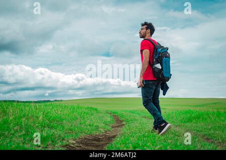 Seitenansicht eines Rucksacktouristen auf einer wunderschönen Straße auf dem Land. Junger Forscher auf einem grünen Feld, Backpacker in einem Stockfoto