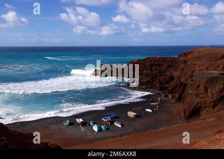 Charco de los Clicos, rote Lavafelsen, Brandung, Boote an Land, schwarzer Lavastrande, Blauer Himmel mit weißen Wolken, Westküste, Lanzarote, Kanarische Inseln, Spanien Stockfoto