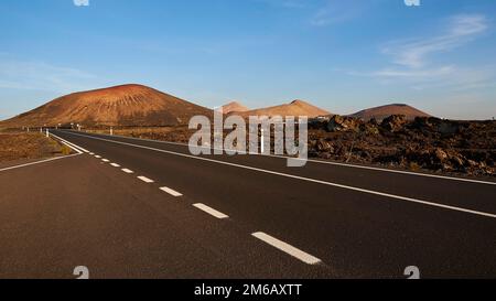 Los vulcanos, Lavalandschaften mit isolierten Vulkanen, rot-braune Erde, schwarze Asphaltstraße ohne Autos, blauer Himmel, wenige weiße Wolken, Lanzarote Stockfoto