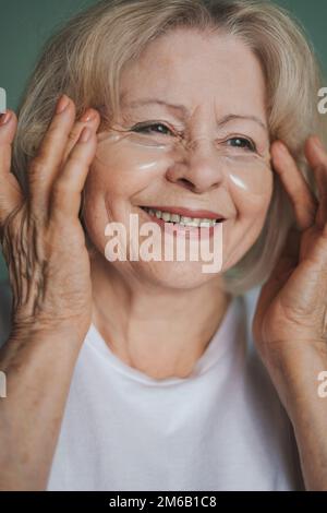 Wunderschöne ältere Frau, die Kollagen-Augenklappen mit Händen hält, isoliert auf grünem Hintergrund. Hautpflegebehandlung Stockfoto