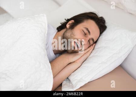 Müde, junger, weißer Mann mit offenem Mund, der in der Freizeit auf einem weißen, weichen Bett im Schlafzimmer schläft, genießen Sie die Ruhe Stockfoto