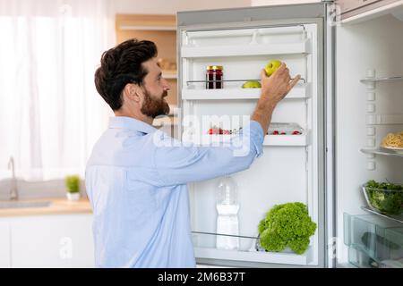 Ein glücklicher hungriger weißer Bärtiger Typ öffnet die Kühlschranktür und nimmt grünen Apfel zum Kochen Stockfoto