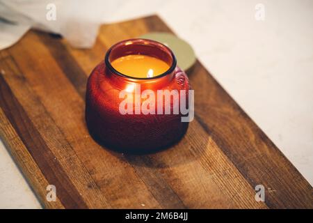 Eine Aromakerze in einem roten Glasgefäß auf einem Holzbrett. Stockfoto