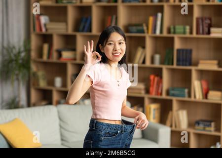 Glückliche asiatische Dame zeigt das Ergebnis von erfolgreichem Gewichtsverlust, Gesten okay und trägt alte übergroße Jeans Stockfoto