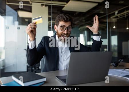 Ein überfreulicher Geschäftsmann mittleren Alters mit offenem Mund, schreiend, mit einer Kreditkarte und einem Laptop im Büro Stockfoto