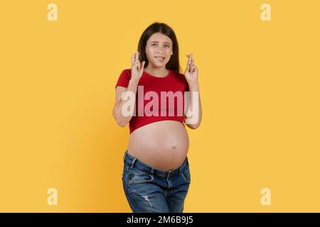 Schwangere Frau, Die Sich Mit Gedrückten Fingern Über Dem Hintergrund Des Yellow Studio Wünscht Stockfoto