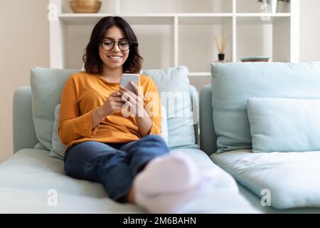 Wunderschöne Arabische Frau Mit Brille, Die Sich Auf Dem Sofa Mit Smartphone Entspannen Kann Stockfoto