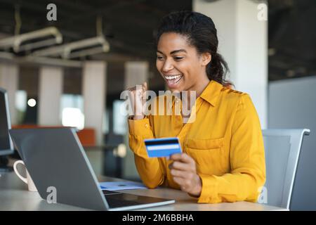 Eine überfreuliche afroamerikanische Geschäftsfrau, die eine erfolgreiche Geste macht, eine Kreditkarte in der Hand hält, mit einem Laptop am Tisch sitzt Stockfoto