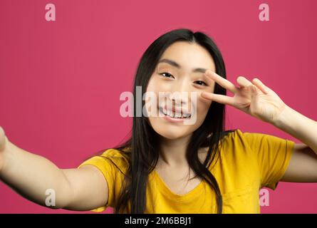 Porträt einer fröhlichen chinesischen Dame, die Selfie macht, Blickwinkel-POV-Aufnahme einer Frau, die in die Kamera schaut und ein V-Zeichen zeigt Stockfoto