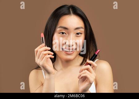 Eine wunderschöne asiatische Frau, die zwei Lippenstifte in der Hand hält und lächelt, und über braunem Studiohintergrund posiert Stockfoto
