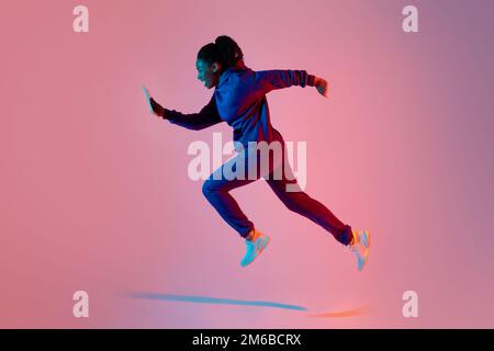 Ganzkörperaufnahme einer modernen, sportlichen Dame, die mit dem Handy springt oder läuft, mit pinkfarbenem Neonhintergrund, Seitenansicht Stockfoto