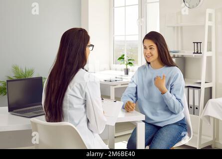 Glückliche junge Patientin erzählt dem Arzt während des Gesprächs in der Arztpraxis von ihrem Gesundheitszustand. Stockfoto