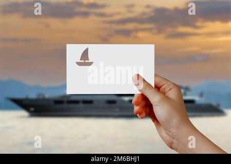 Die Hand eines Mannes, der das Yachtsymbol vor dem Meer hält. Konzept von Reise, Reise, Traum, Freiheit. Hand hält Papierboot gegen Yachtstation wi Stockfoto