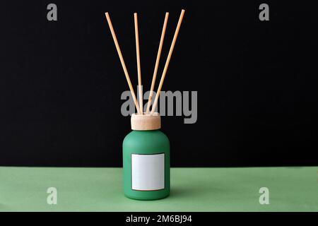 Reed-Diffusor-Ölflasche mit leerem weißem Etikett auf grünem Sockel, isoliert vor schwarzem Hintergrund. Speicherplatz kopieren. Keine Menschen. Stockfoto