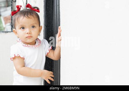 Wunderschönes braungehäutetes Latina-Baby, das neben einer weißen Wand steht und vor Neugier und Angst aussieht. Stockfoto