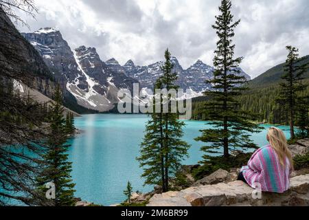 Eine abenteuerlustige blonde Frau sitzt am Rande einer Klippe am Moraine Lake und bewundert die Kanadischen Rocky Mountains Stockfoto