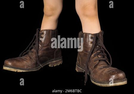 Ein Kind, das ein Paar zu große, ungebundene und unpolierte braune Lederstiefel trägt Stockfoto