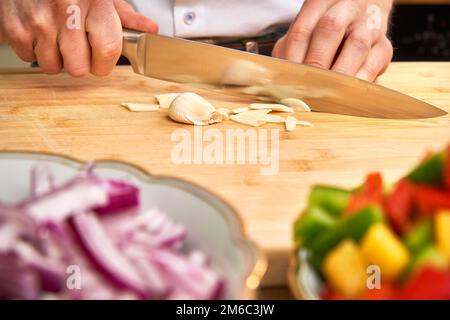 Hände des Mannes Schneiden von frischem Knoblauch in der Küche eine Mahlzeit zuzubereiten, für das Mittagessen. Paprika und Zwiebeln im Vordergrund. Stockfoto