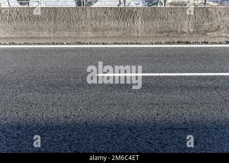 Asphalt einer Straße mit weißer Farbe, die die Spuren trennt, und einer Betonwand, die die Fahrwege trennt Stockfoto