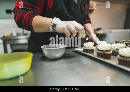 Ein Bäcker mit rotem Karohemd, schwarzer Schürze und transparenten Handschuhen, der Cupcakes mit weißer Sahne dekoriert, die auf ein Backblech in der Küche gelegt wird. Hochwertiges Foto Stockfoto