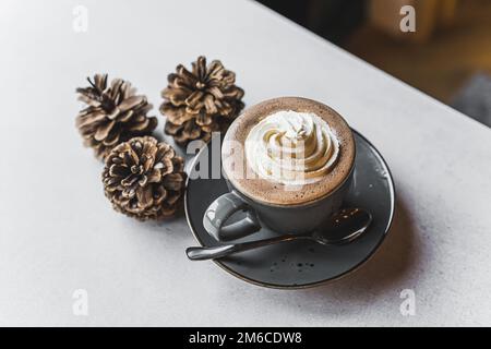 Kaffee dekoriert mit Schlagsahne in einer grauen Tasse auf einem grauen Teller mit einem Löffel. Drei Kegel neben dem Lagerring. Hochwertiges Foto Stockfoto
