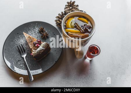 Nachtisch und ein heißes Getränk. Ein Stück Kuchen mit Puderzucker und Früchten auf einem schwarzen Teller neben einer weißen Tasse mit einem warmen winterlichen Getränk und einem kleinen Glas rotem Sirup. Hochwertiges Foto Stockfoto