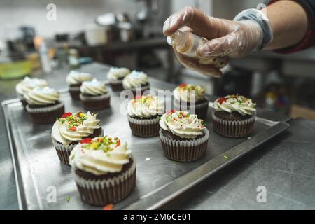 Cupcakes dekoriert mit weißer Creme und farbenfrohen Glitzern, die in der Küche von einer Person zubereitet werden. Hand in einen transparenten Handschuh. Unscharfer Hintergrund. Hochwertiges Foto Stockfoto