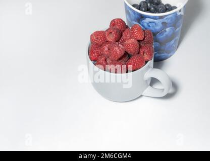 Himbeeren in einem großen Becher und Blaubeeren und Pappbecher auf weißem Hintergrund Stockfoto