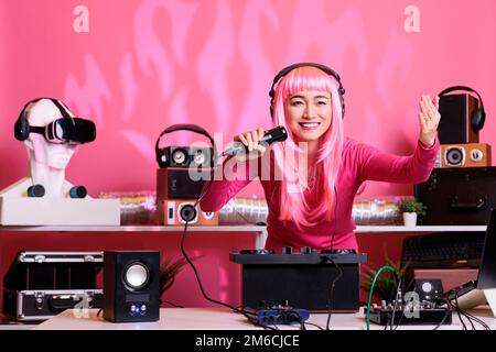 Asiatischer Musiker sitzt am dj-Tisch und spielt Techno-Musik auf professionellen Plattenspielern, trägt ein Headset und spricht mit Fans über das Mikrofon. Künstler mit pinkfarbenen Haaren, der nachts ein elektronisches Lied spielt Stockfoto